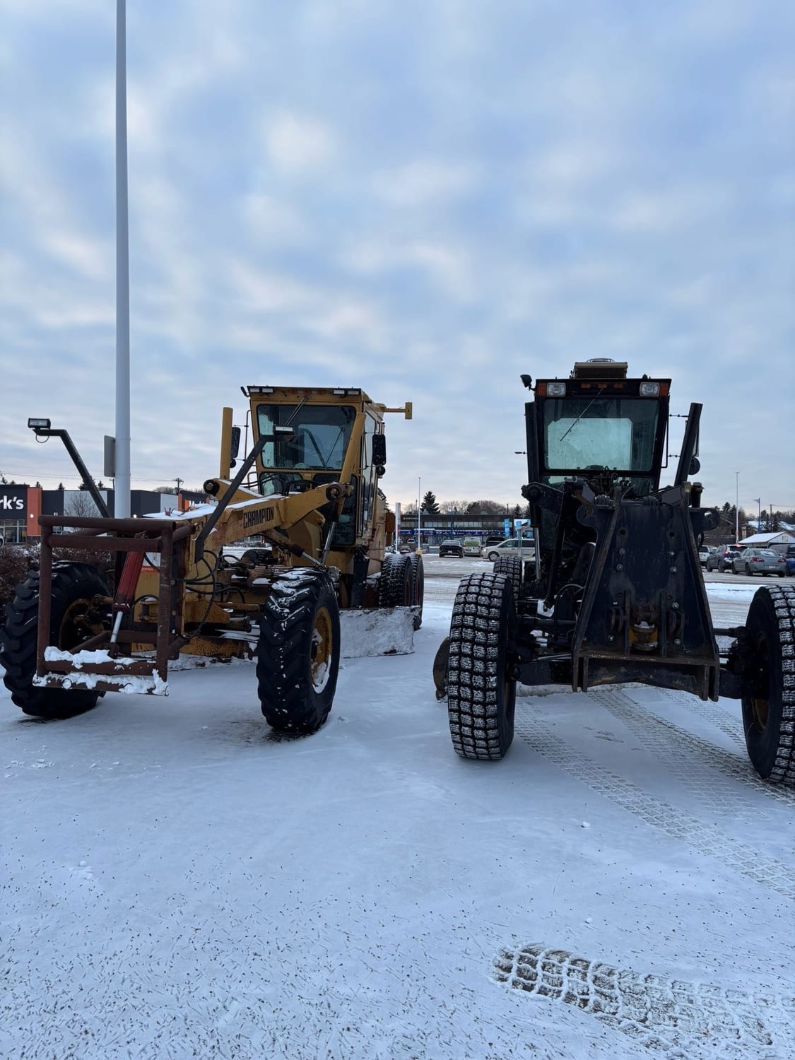 Two motor graders with plow blades on a snow-covered parking lot