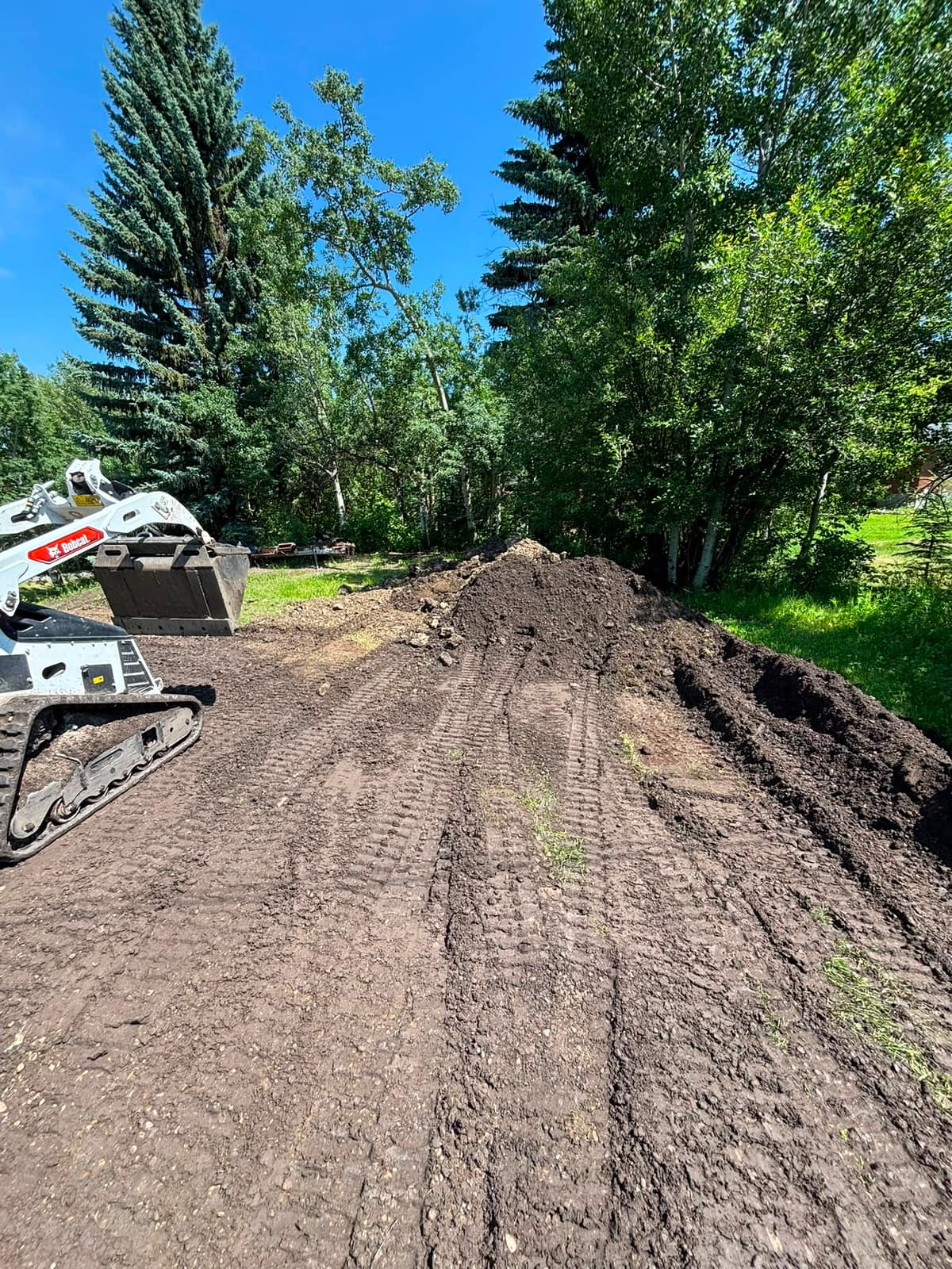 Bobcat track loader spreading topsoil on a treed acreage